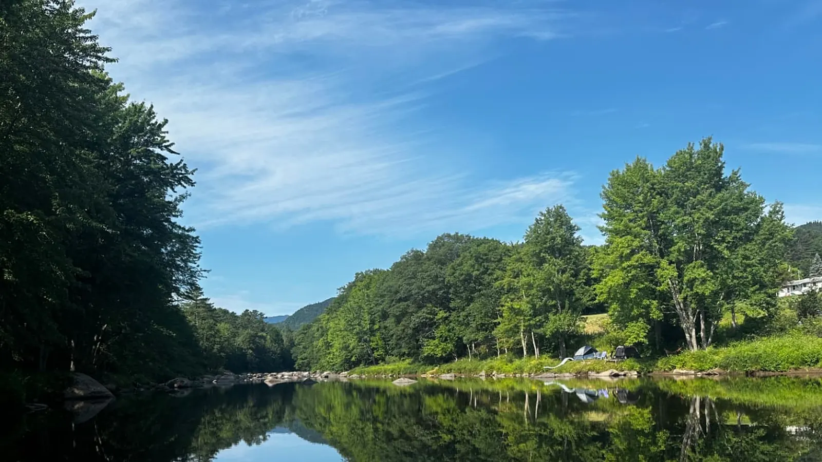A calm bend of the AuSable River mirroring the trees on both banks, with ledge rock in the clear shallows in the foreground